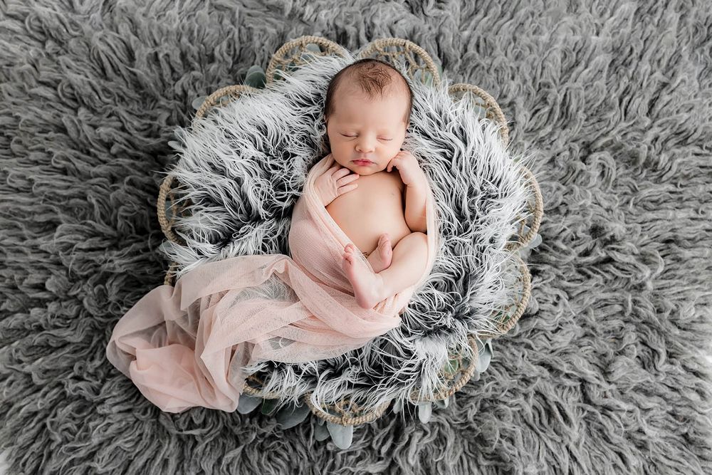 Sleeping newborn baby wrapped in pink swaddle nestled in a basket on gray fluffy rug.