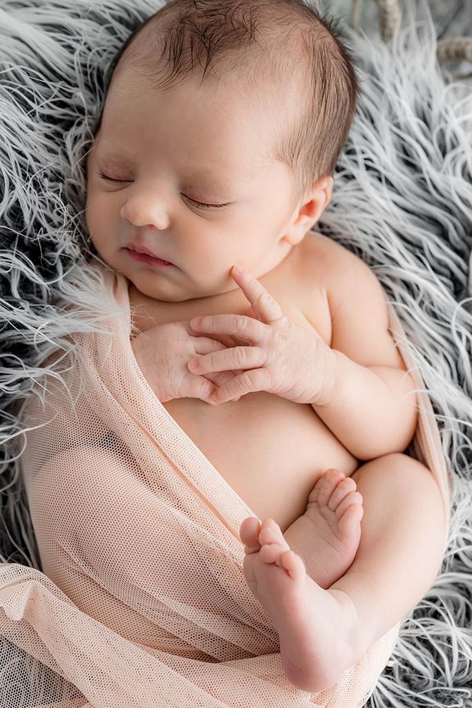 Peaceful newborn baby sleeping on a fluffy gray blanket, wrapped in a soft pink cloth with tiny hands and feet visible.