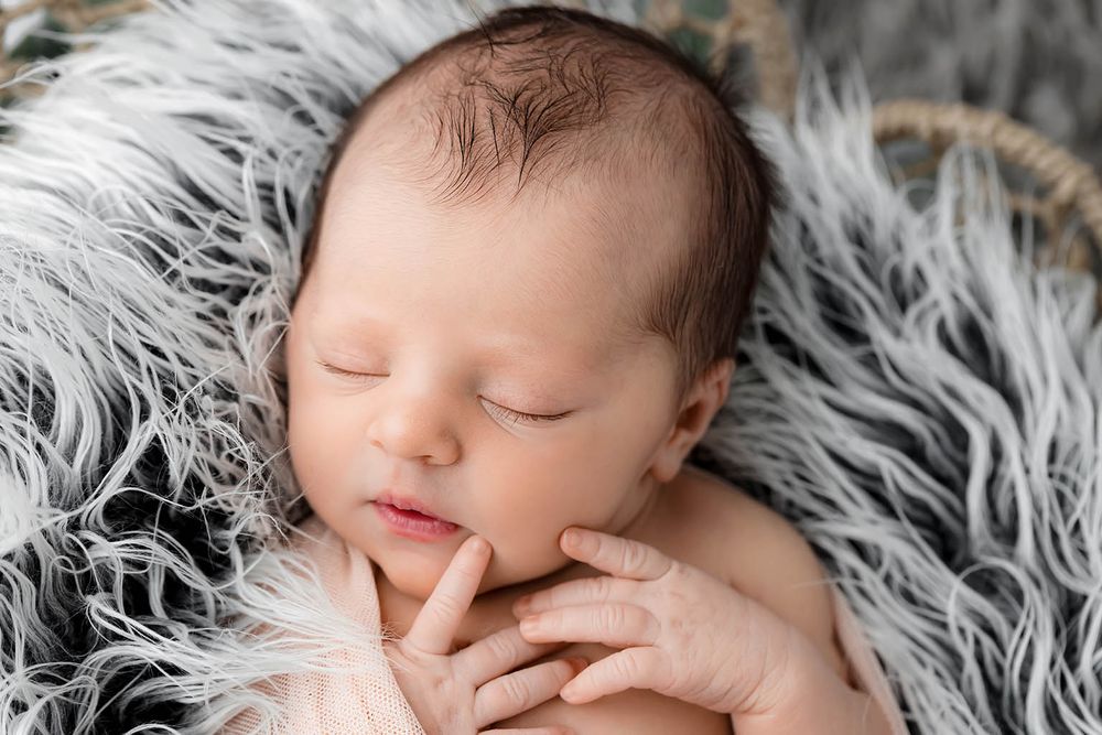 Close-up of peaceful sleeping newborn baby with hands tucked under chin on fluffy gray fur.