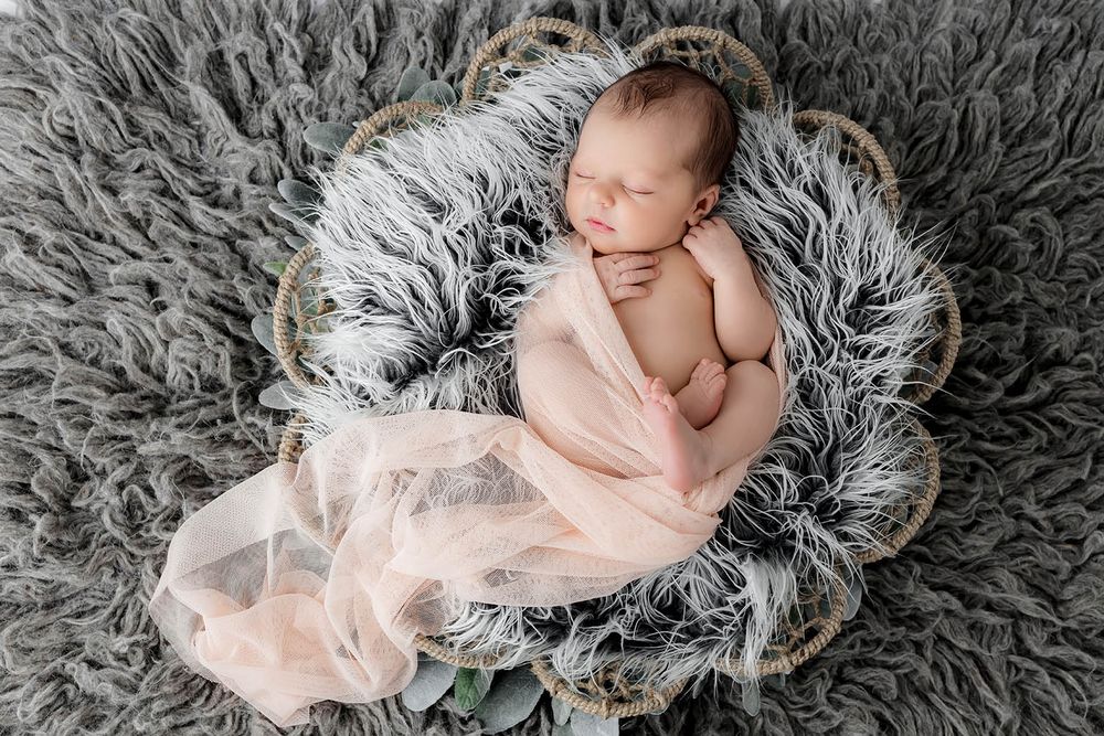 Sleeping newborn baby wrapped in pink fabric in a round wicker basket on a gray fluffy rug.