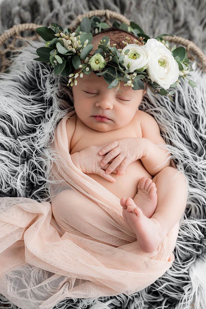 Sleeping newborn baby wearing floral crown wrapped in pink cloth in a basket on gray rug.