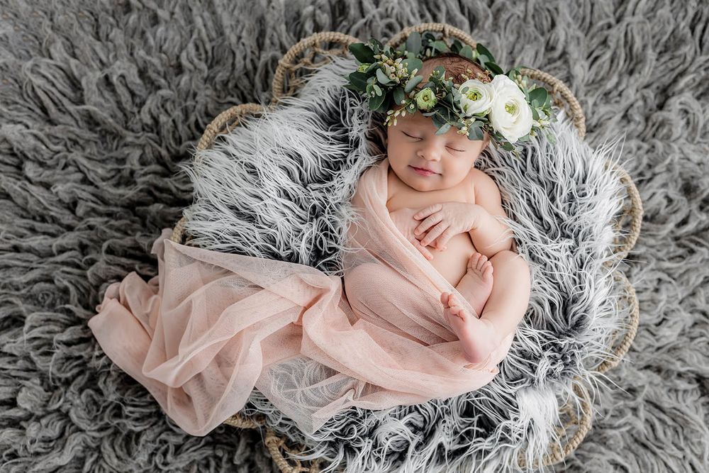Sleeping newborn baby girl wrapped in pink cloth with floral crown in wicker basket on gray fur.