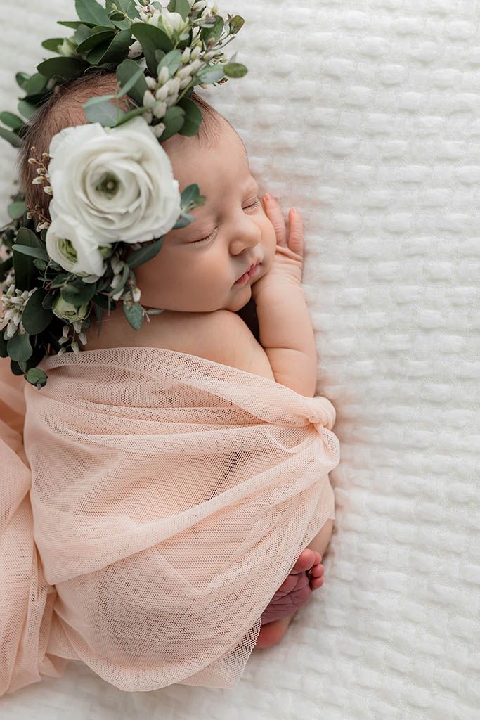Peaceful newborn baby girl sleeping wrapped in blush pink fabric with white flower and greenery crown.