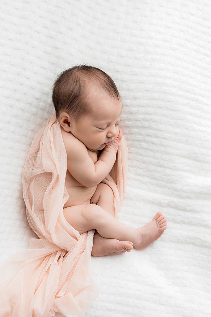 Sleeping newborn baby wrapped in a soft peach fabric on a white knit blanket.