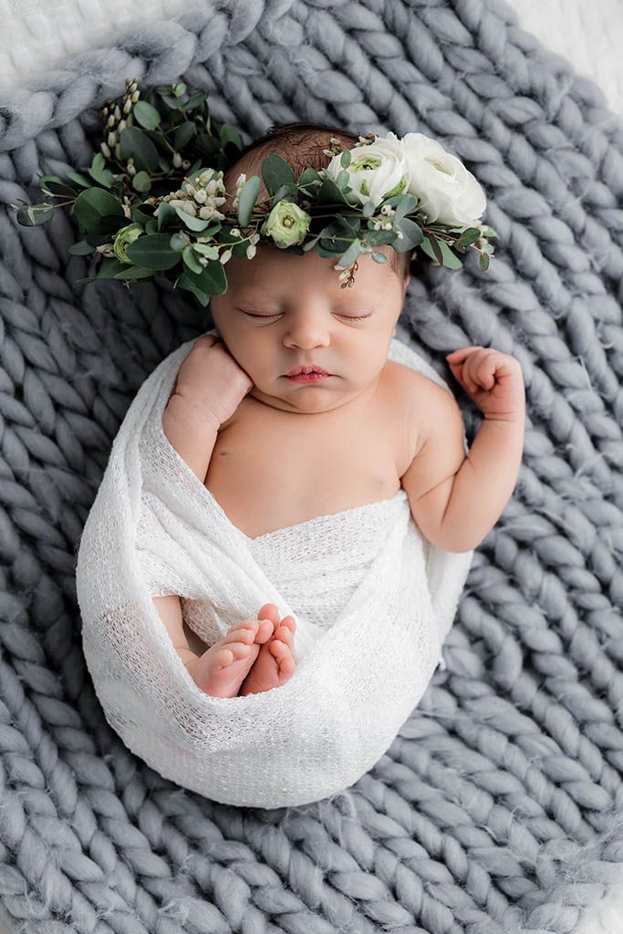 Newborn baby swaddled in white wrap wearing floral crown resting on gray knit blanket.