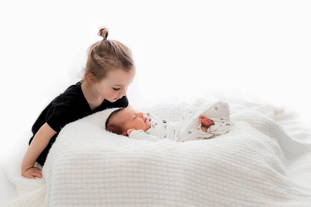 Young girl lovingly looks at newborn sibling wrapped in white blanket during studio photo session.