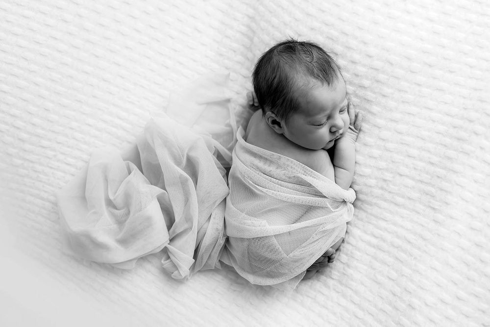 Black and white photo of sleeping newborn baby wrapped in sheer fabric on white blanket.