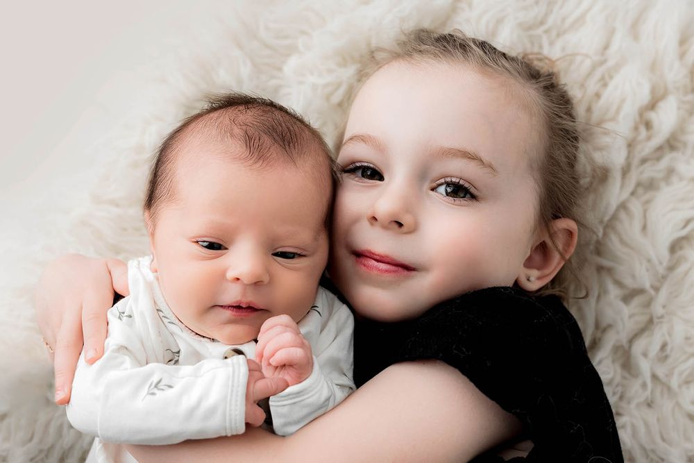 Smiling young girl with blue eyes gently holding a newborn baby sibling on a white fluffy blanket.