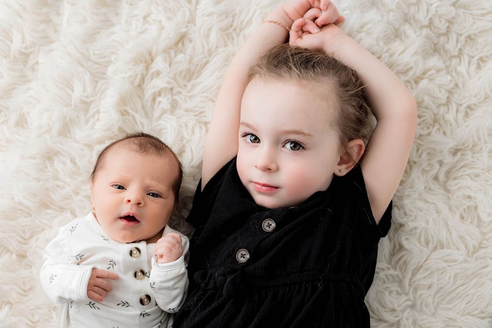 Newborn baby and toddler sibling lying together on a white fluffy blanket.