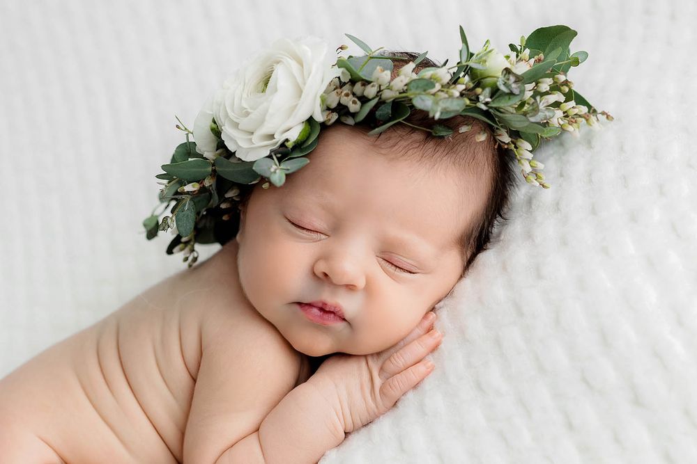 Sleeping newborn baby wearing a white floral crown with greenery on a white textured blanket.