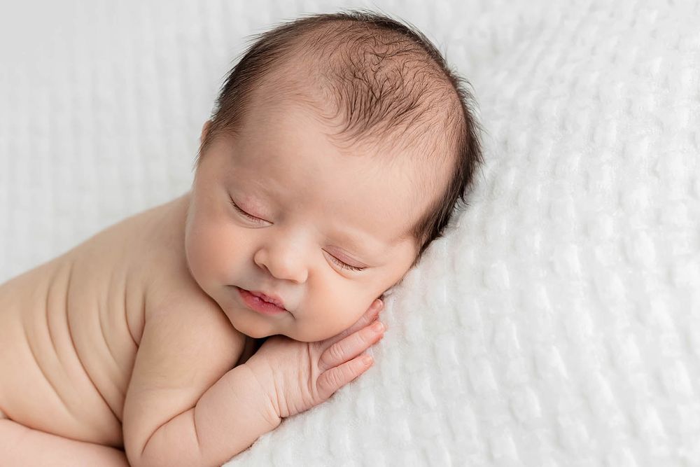 Peaceful sleeping newborn baby resting on white textured blanket with hands under cheek.