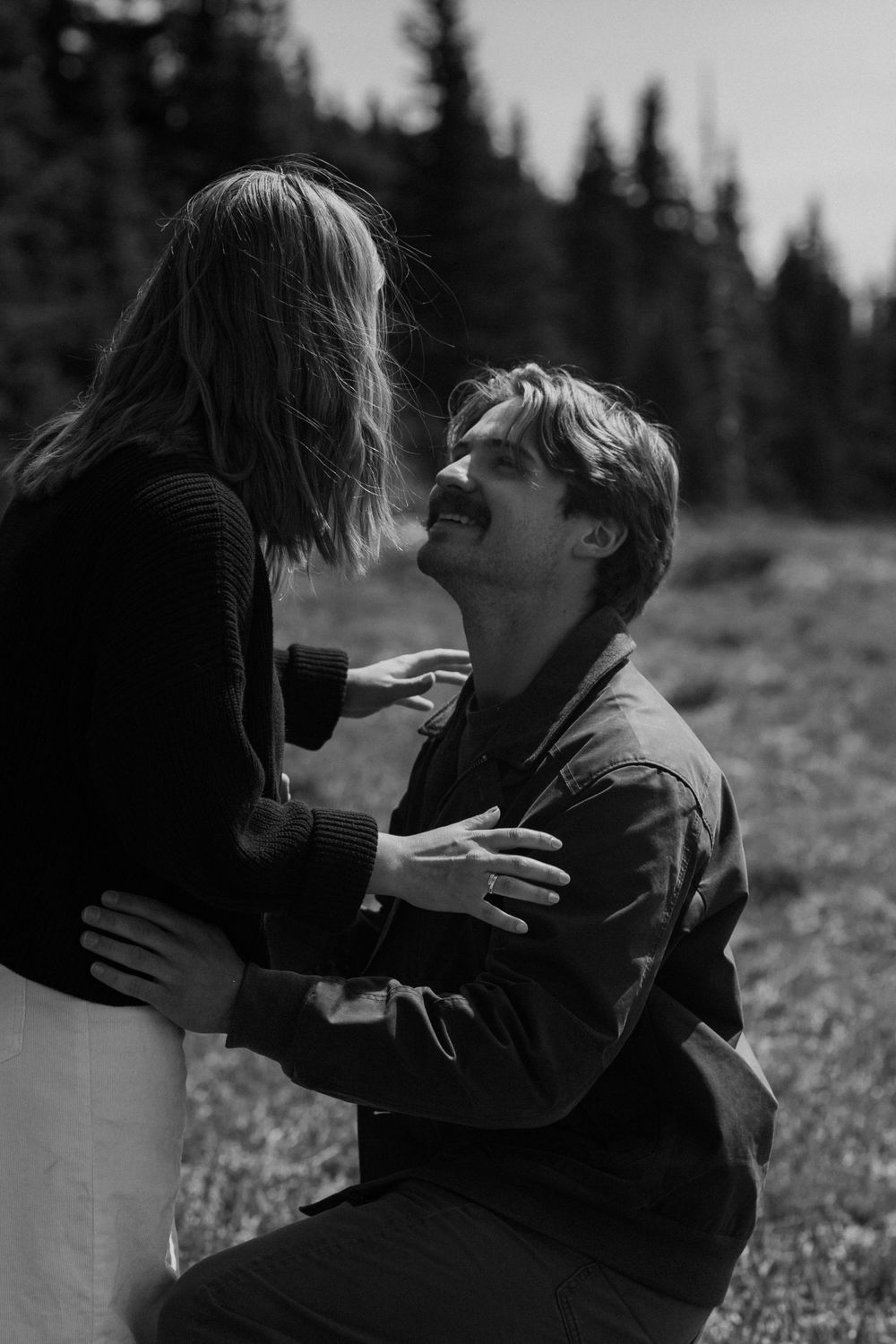 Black and white photo of a man kneeling and smiling up at a woman outdoors during a romantic proposal.