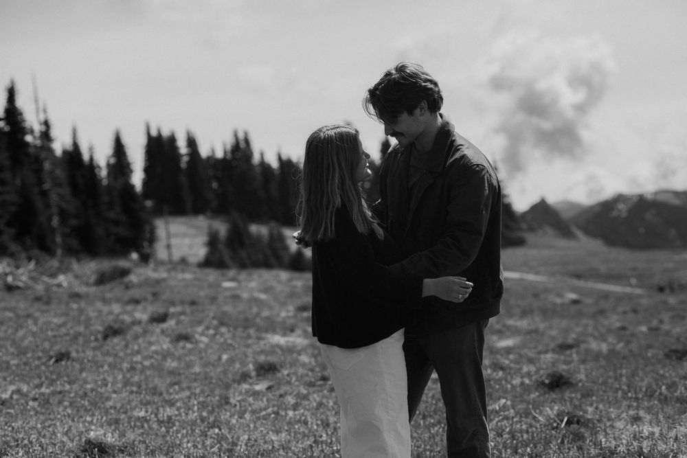 Black and white photo of a couple sharing an intimate moment in a mountain meadow surrounded by evergreen trees.