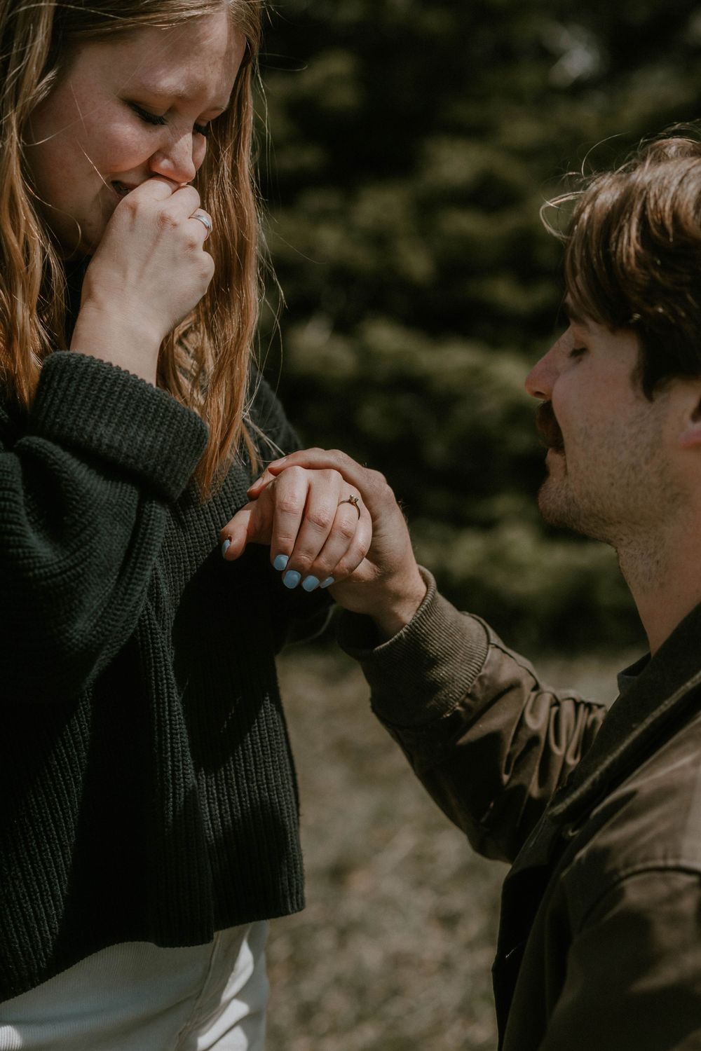 Man kneeling to propose to emotional woman outdoors, holding her hand near evergreen trees.