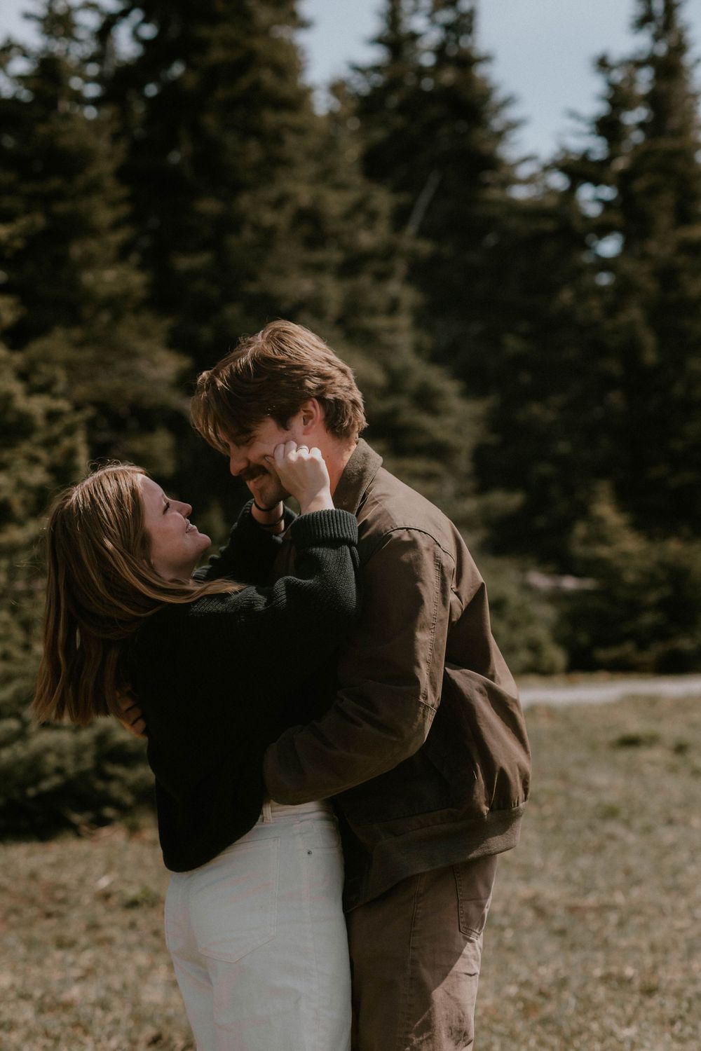 A couple embraces and laughs together outdoors with evergreen trees in the background.