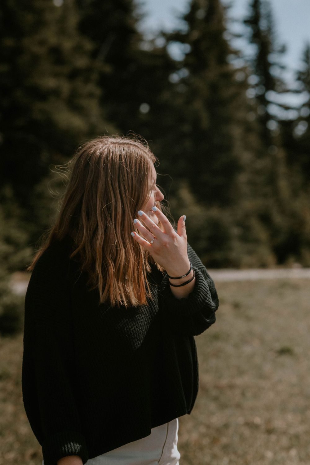 Woman in black sweater standing outdoors near pine trees, hand raised near face in natural forest setting.