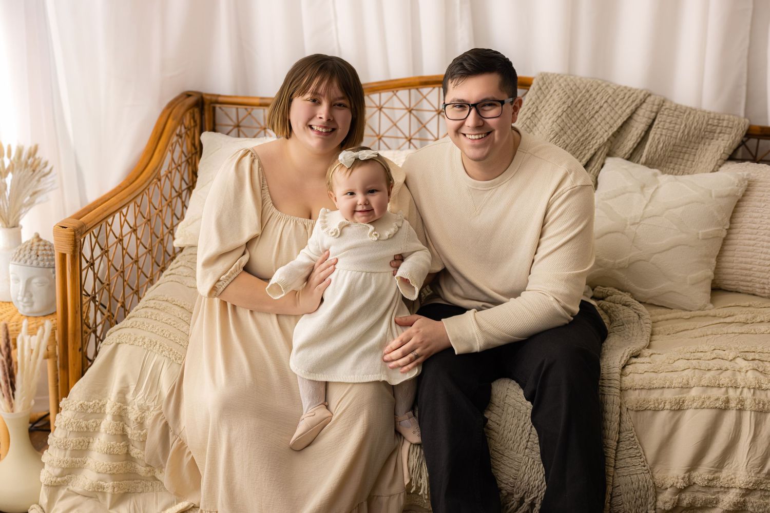 A happy family of 3 sitting on a rattan daybed with neutral coverings, smiling at the camera