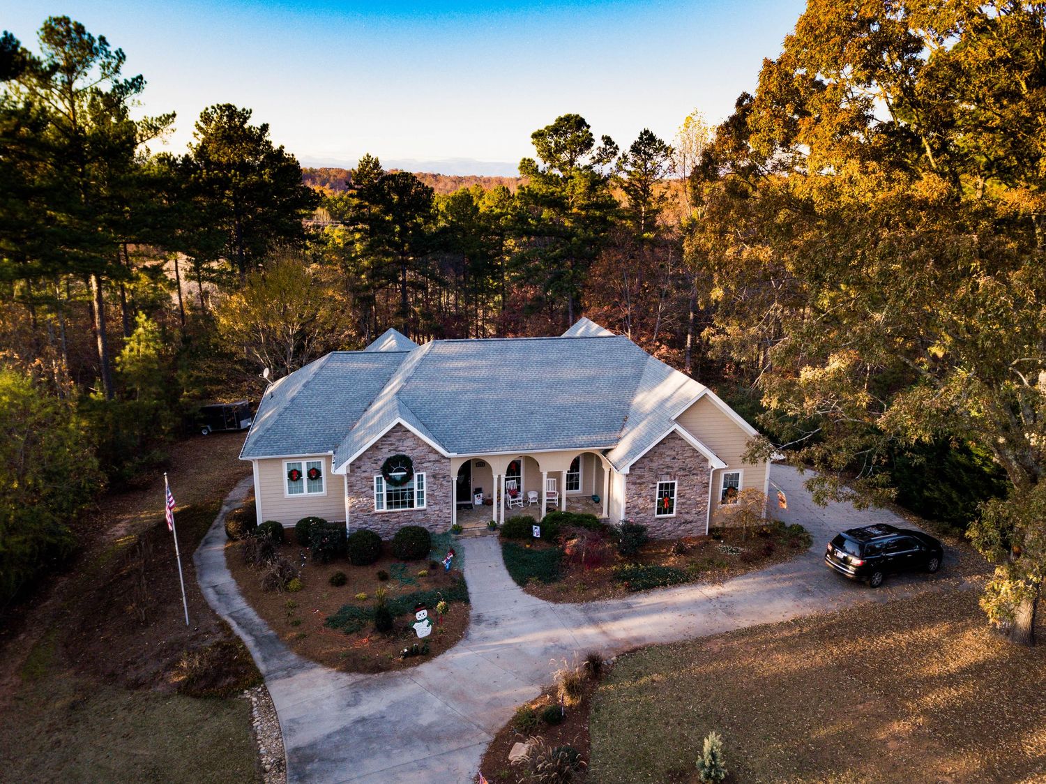 Aerial view of a brick ranch home surrounded by mature trees with a circular driveway and parked car at golden hour.