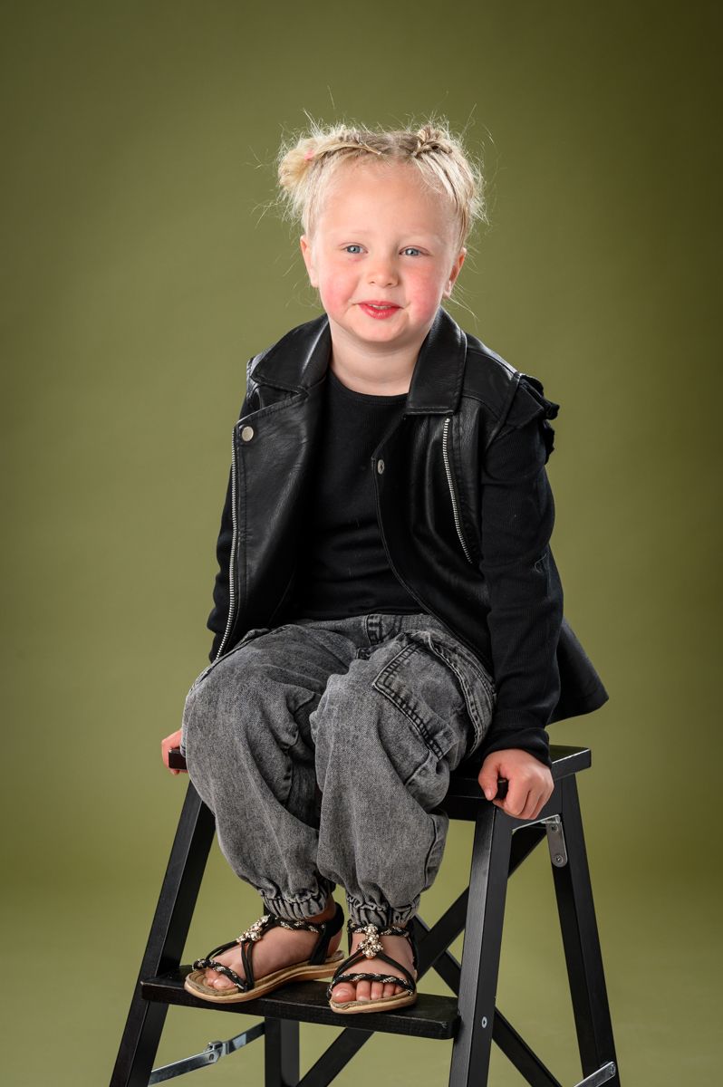 Child wearing black jacket and jeans sitting on a wooden chair against olive green background.