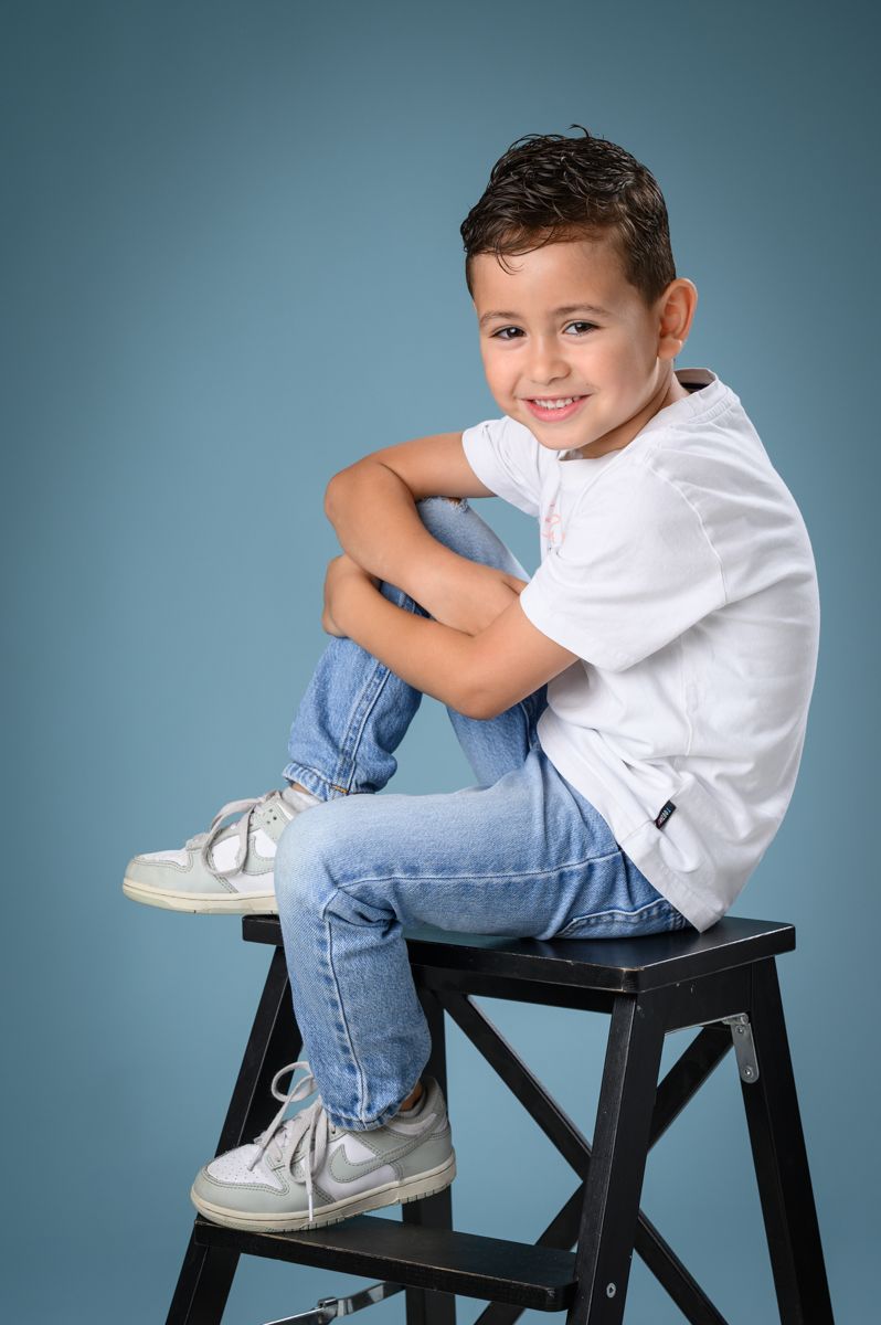 Child in white t-shirt and jeans sitting casually on black chair against blue background.