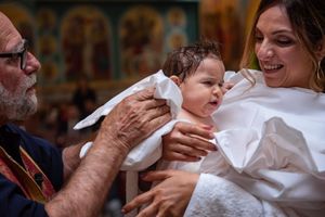 Smiling priest and mother hold a baby wrapped in white baptismal cloth during an Orthodox christening.