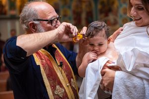 Orthodox priest anoints a baby with holy oil during a christening ceremony while mother looks on smiling.