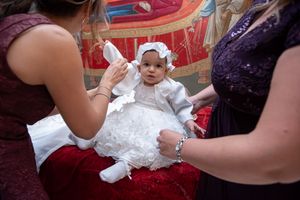 Baby dressed in white christening gown sits on red velvet cushion during Orthodox baptism ceremony.