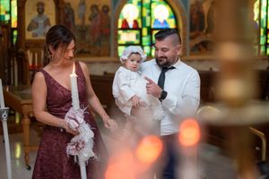 Family stands together holding baptismal candle with baby in white christening outfit inside Orthodox church.