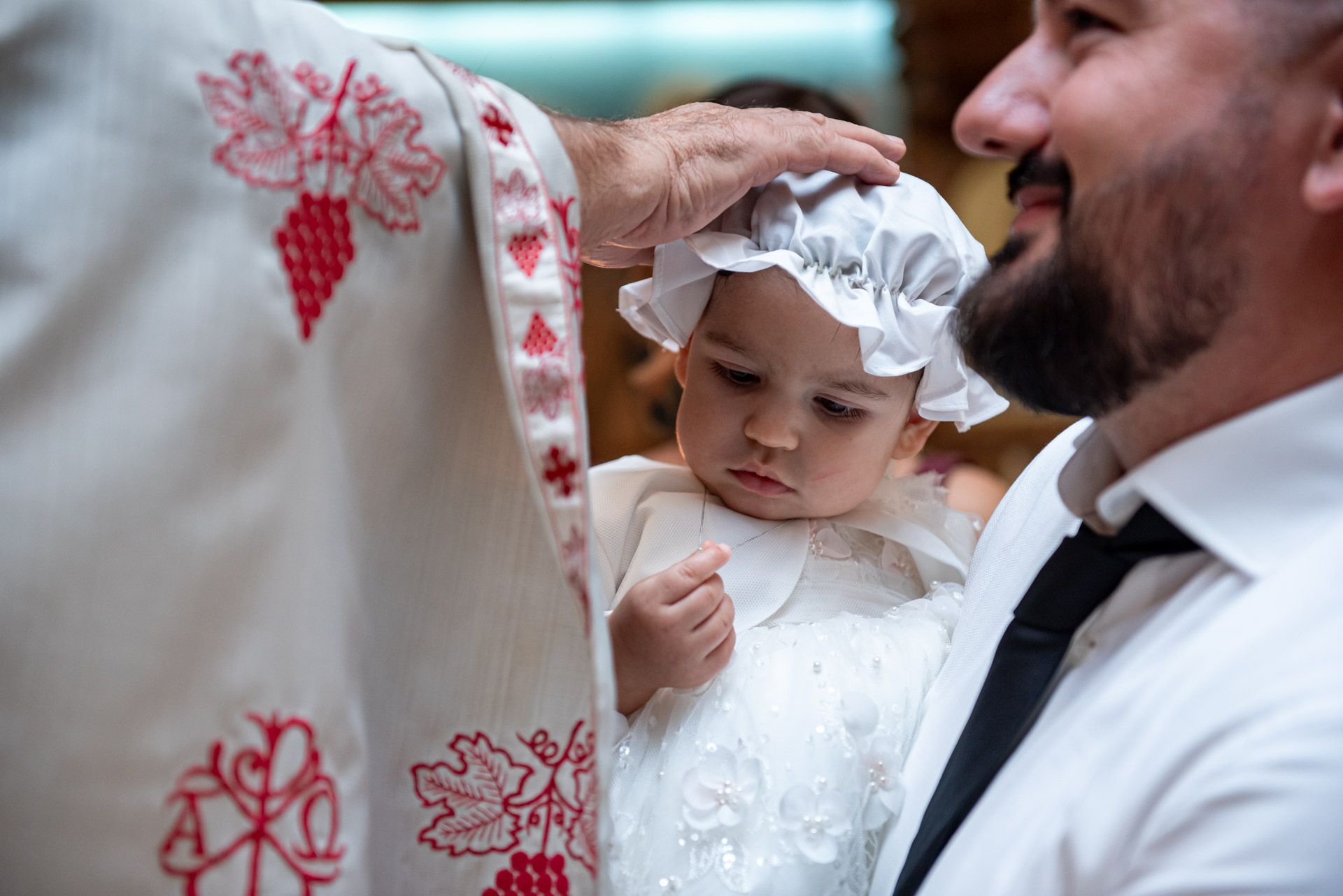 Orthodox priest places hand on baby wearing white bonnet during christening rite held by smiling father.