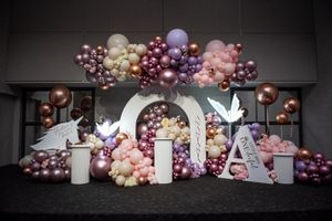 Elegant baptism party backdrop with pink purple and gold balloon garland arch and illuminated letter decorations.