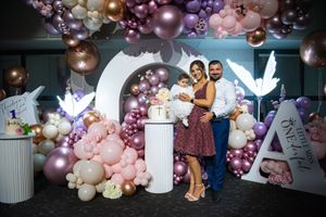 Family poses with baby at first birthday celebration in front of pink and purple balloon arch display.
