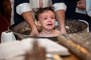 A crying toddler sitting in an ornate silver baptismal font during a baptism ceremony.