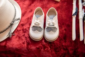 White baby baptism shoes with bow ties displayed on red velvet beside a hat.