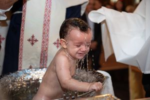 A crying baby being baptized with water poured over them in a decorative baptismal font.
