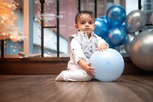 A baby boy in white outfit sitting on a wooden floor holding a light blue balloon.