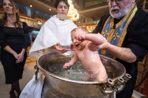 A crying baby being baptized by immersion in a large font by an Orthodox priest during a christening ceremony.