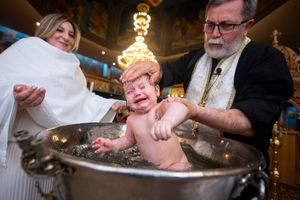 Orthodox priest baptizing crying baby by immersion in silver font during christening ceremony.