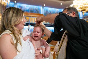 Orthodox priest anointing crying baby held by godmother during christening ceremony in church.