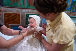 Baby girl in white lace baptism gown and bonnet being dressed for christening ceremony.