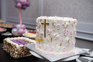 White baptism cake decorated with gold cross, pink flowers, and name topper at christening reception.