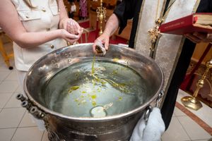 Orthodox priest blessing water with oil in large silver baptismal font during christening.