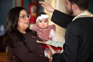 Orthodox priest blessing baby girl held by grandmother during baptism ceremony.