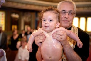 Smiling baby held by Orthodox priest during a church baptism ceremony.