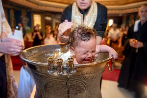 Crying baby being immersed in baptismal font during Orthodox Christian christening.