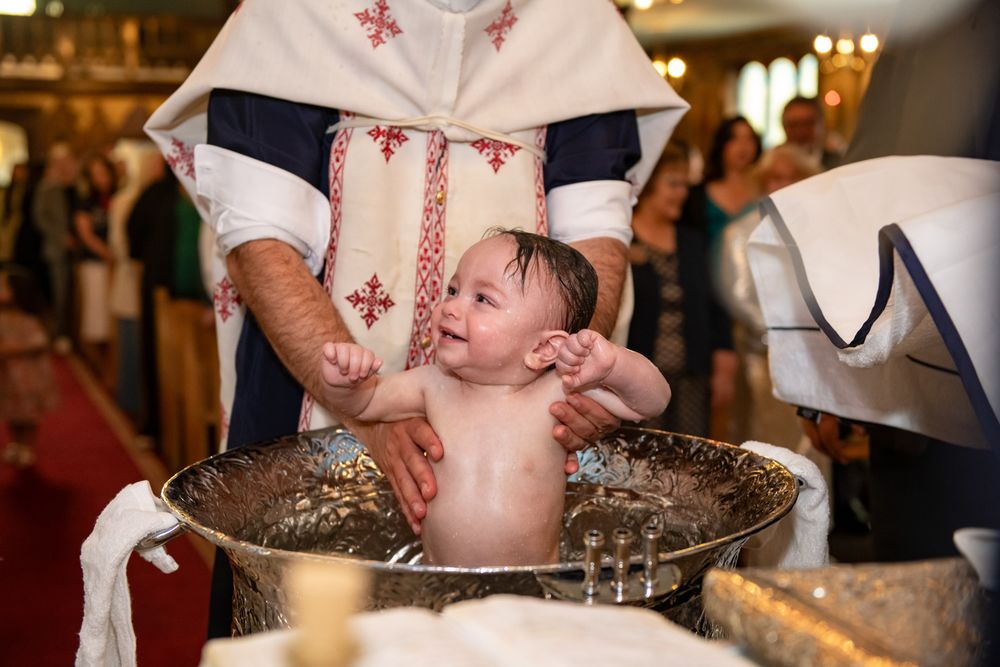 Smiling baby being held in a golden baptismal font by an Orthodox priest during christening ceremony.