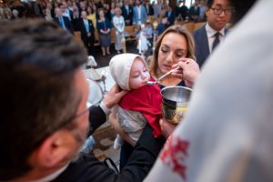 Baby in white baptism cap receiving Holy Communion from a priest during an Orthodox christening service.