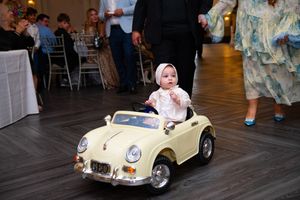 A baby in a white outfit rides a vintage yellow toy car on a dance floor at an elegant event.
