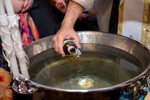 Olive oil being poured into a large silver baptismal font during an Orthodox Christian baptism ceremony.