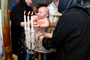 A baby being immersed in water during an Orthodox Christian baptism with candles and robed attendants.