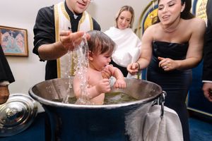 A baby being baptized in a large metal font with water being poured over them by a priest.